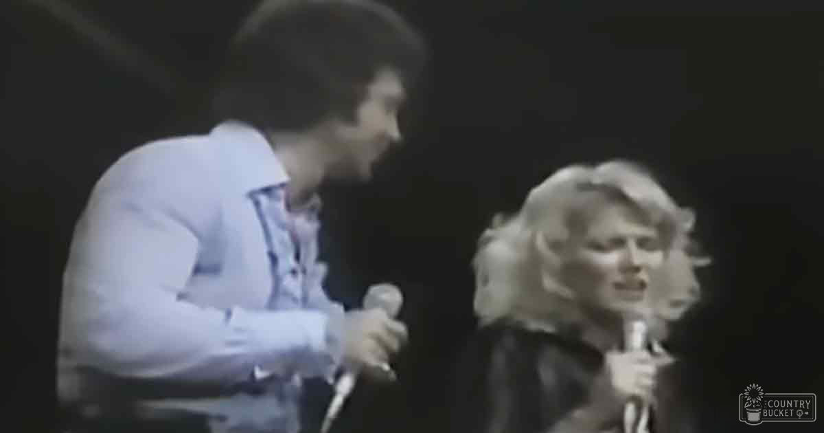 A young adult male singer in a light-colored shirt performs on stage with an adult female singer, both holding microphones and wearing dark outfits against a dimly lit background.