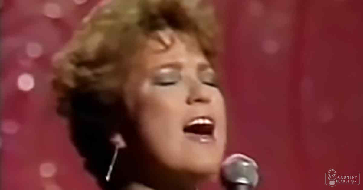 Adult female singer wearing a fitted dark red or maroon blouse with earrings, singing into a handheld silver microphone on stage with circular light patterns against a red curtain backdrop at night.
