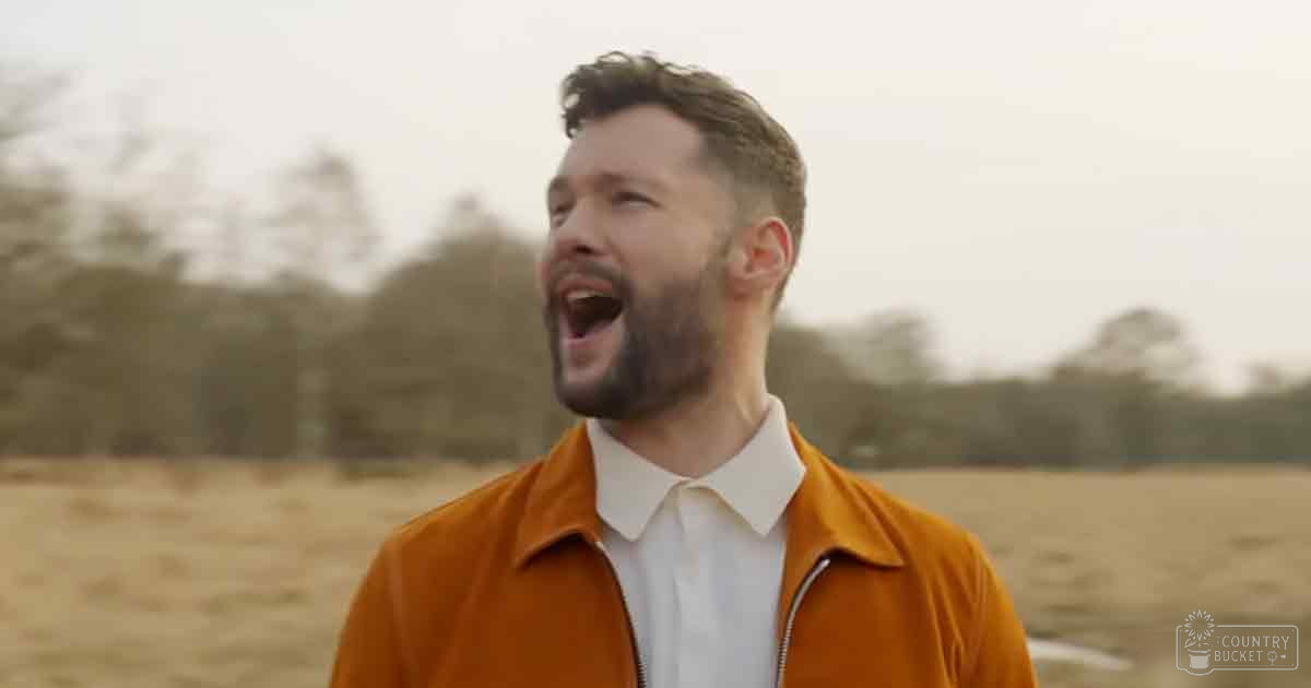 A young adult male wearing an orange jacket over a white collared shirt stands in an open field with bare trees suggesting autumn. The setting is soft and diffused, indicating either an overcast day or late afternoon/early evening.