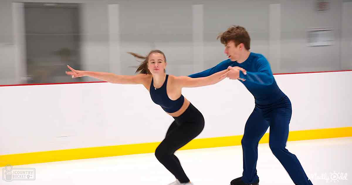 A young adult female figure skater in a dark blue sports bra and black leggings practices alongside an equally youthful male partner at an indoor ice rink with white walls, red and yellow barriers.