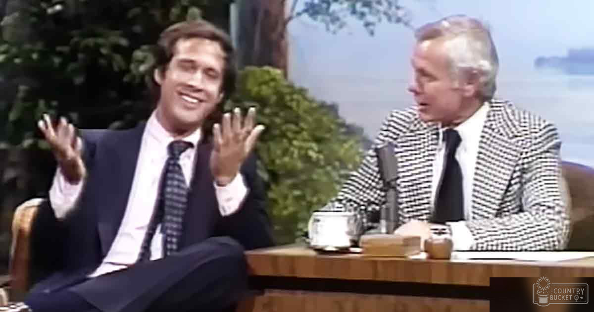 A talk show host in an outdoor setting with two male adults wearing formal attire: one in a dark suit and another in a checkered blazer, surrounded by natural scenery with trees and foliage.