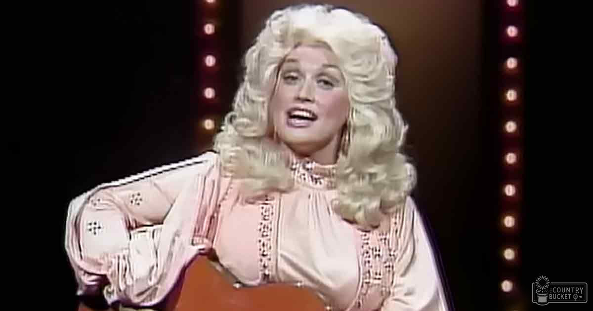 An adult female performer wearing a long-sleeved, embroidered blouse in an indoor performance setting with stage lighting and holding a guitar.