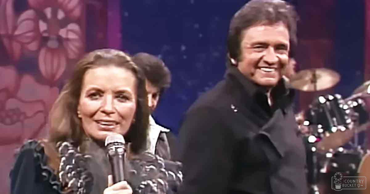 A female performer wearing an intricately patterned dress holds a microphone while standing next to another adult male in a tailored jacket on stage in what appears to be a studio setting with musical instruments and floral backdrop.