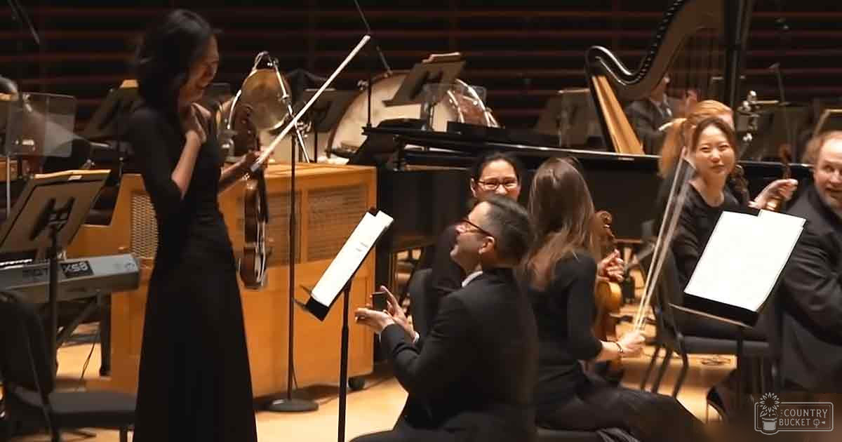 Young adult female singer in a formal orchestra setting during what appears to be a classical music performance, surrounded by seated musicians in a concert hall with wooden paneling and acoustic treatment.