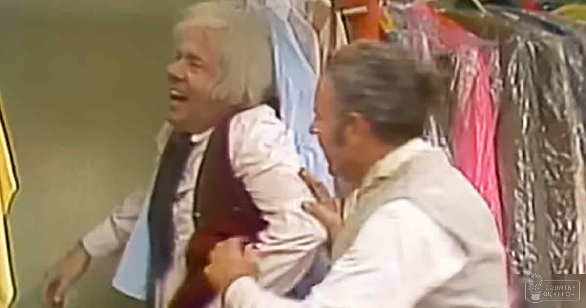 A scene from a theatrical performance or comedy skit set in a dressing room with two adult males wearing different garments and various clothing hanging on racks.