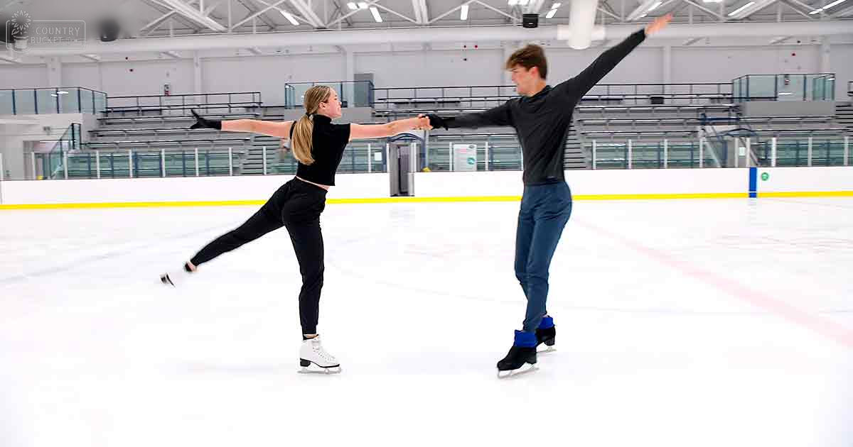 A young couple practicing figure skating in an indoor ice rink with white walls, yellow safety lines, and spectator seating in the background.