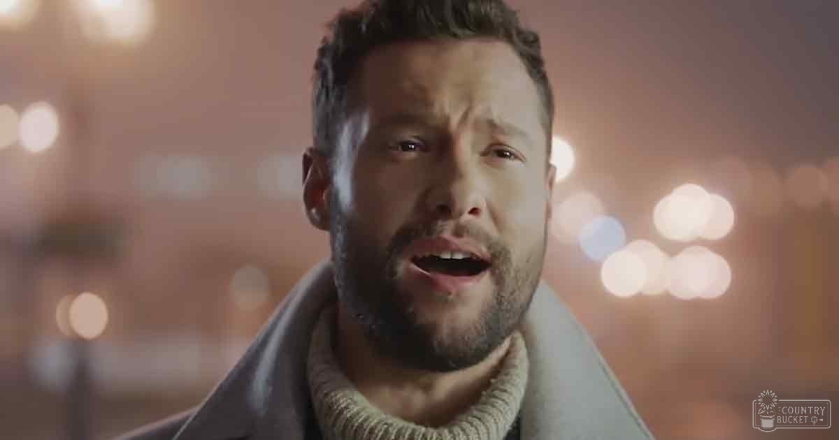 A young adult male in a light-colored turtleneck sweater under a gray coat with a high collar stands in an urban nighttime setting, surrounded by muted tones and warm artificial lighting.