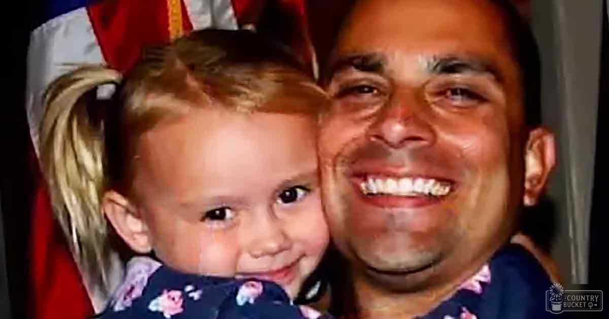 A middle-aged man holds his toddler daughter indoors near an American flag, both wearing dark blue garments with floral patterns.