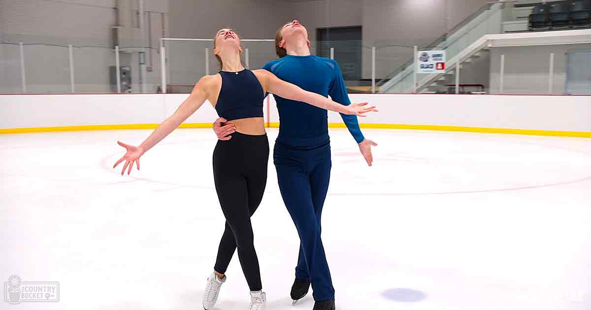 Two young adults practicing figure skating on an indoor ice rink with white walls and yellow barriers.