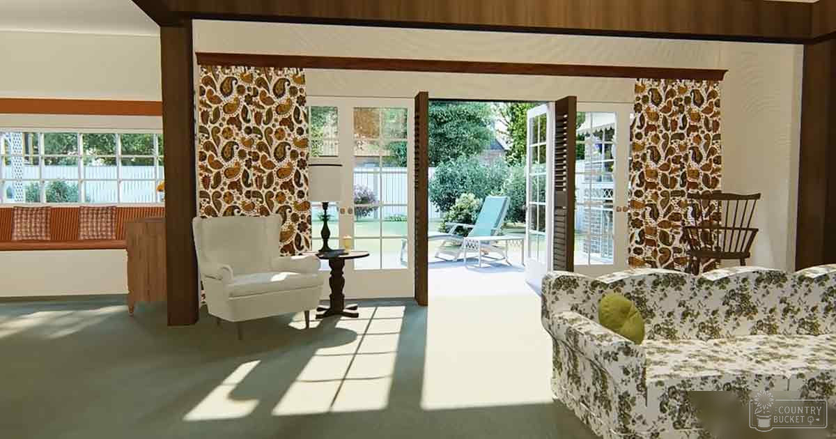 A cozy living room scene with a white armchair, floral-patterned sofa, wooden table, and large windows showing an outdoor garden in daylight.