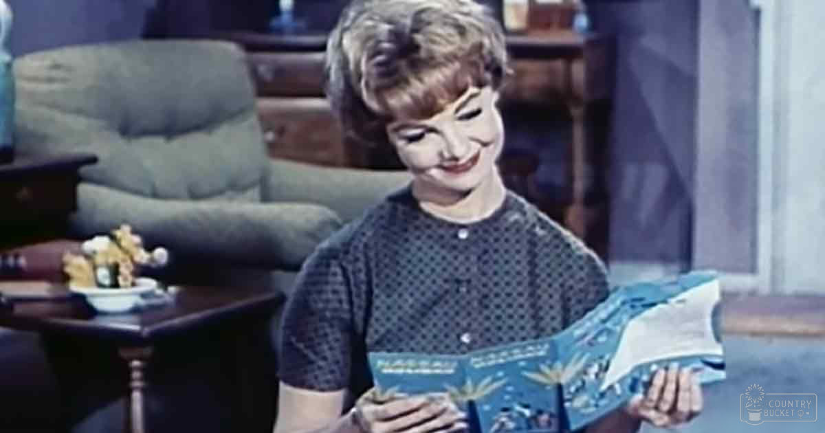 A young adult female in a mid-20th-century indoor setting, wearing a dark blue polka dot dress with cotton fabric, holding an open book.