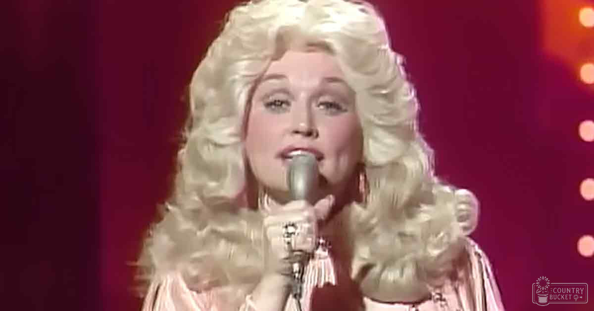 A female performer in a flowy, light pink dress with ruffled details and large hoop earrings is on stage under red lighting at an indoor venue.