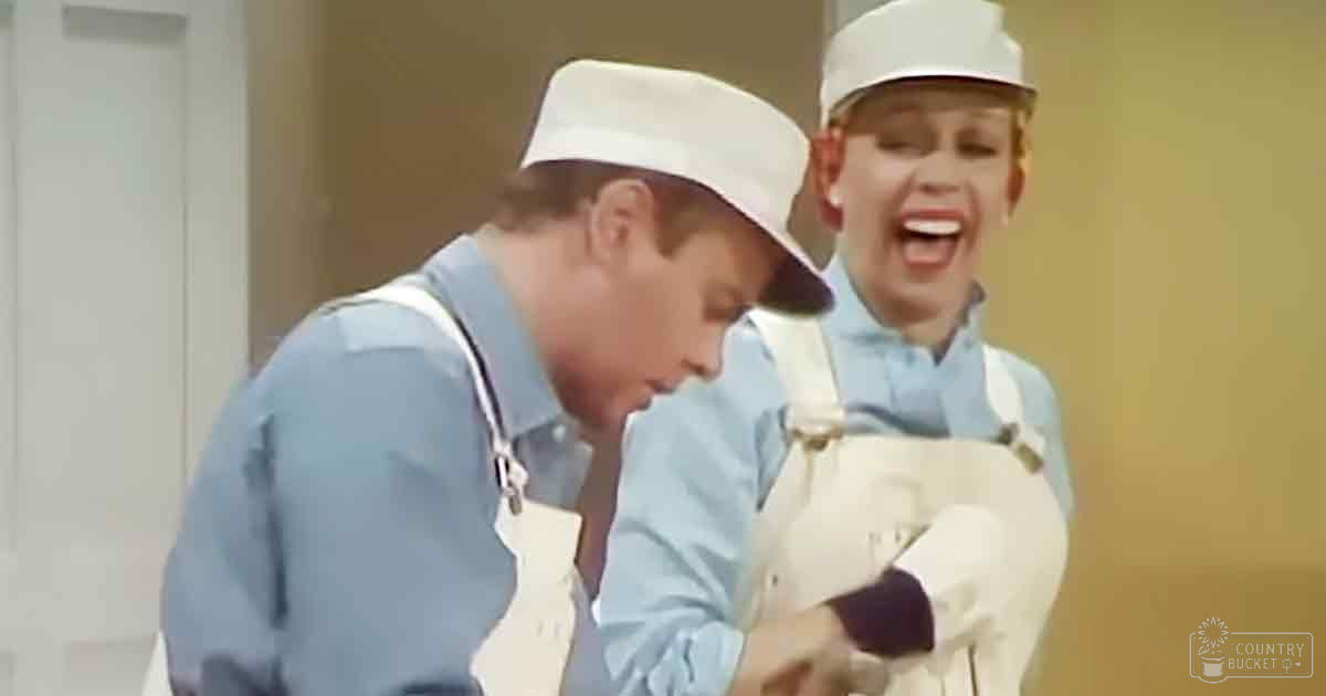 Young adult male in blue shirt with stethoscope and young female child in light blue shirt and white hat, indoor medical setting during daytime or early evening.