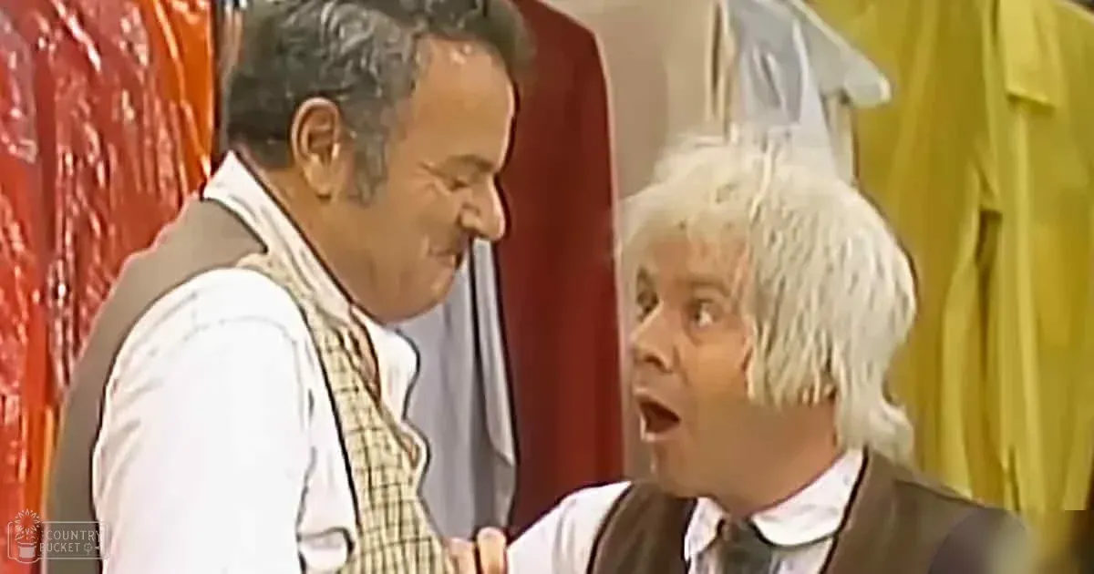 A comedic scene from a television show or film set in an indoor location, with two middle-aged men wearing white shirts with checkered and dark brown vests over them, standing against red and yellow curtains suggesting theatrical elements.