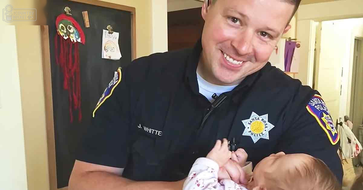 A police officer holding an infant in a home setting with a blackboard and notes visible, natural light suggesting daytime.