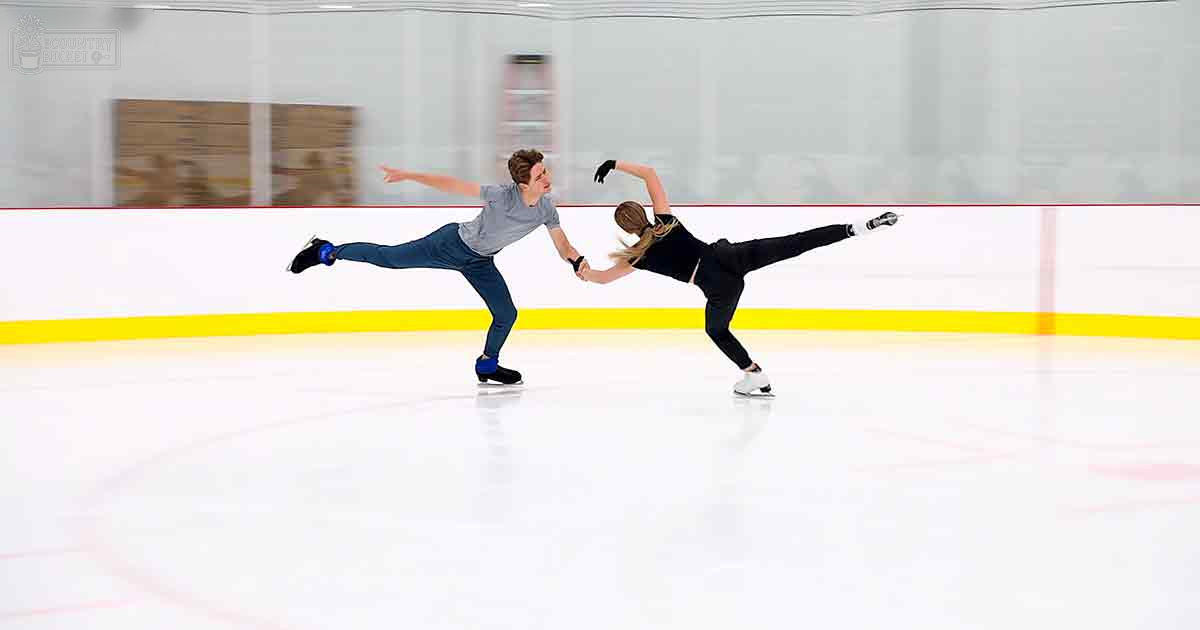 A male figure skater in a gray t-shirt and dark blue jeans, alongside a female skater in a black leotard, skating on an indoor ice rink with yellow line markings and red borders. The scene is well-lit by artificial light.