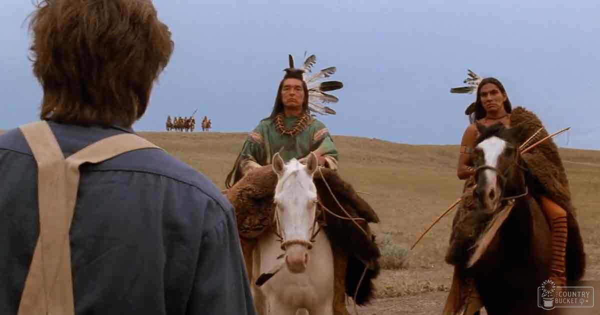 An adult male wearing traditional Native American attire sits on a white horse in an open landscape during what appears to be the Western frontier era.