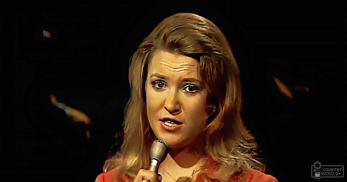 An adult female stands indoors under artificial stage lighting, wearing a relaxed, coral-pink blouse with a rounded neckline.