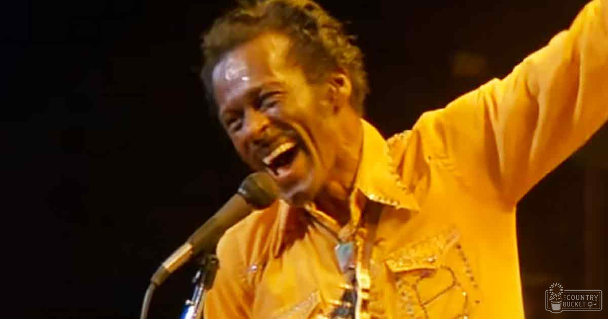 A middle-aged male singer wearing a light brown, decorative shirt with short sleeves sings into a microphone at an event.