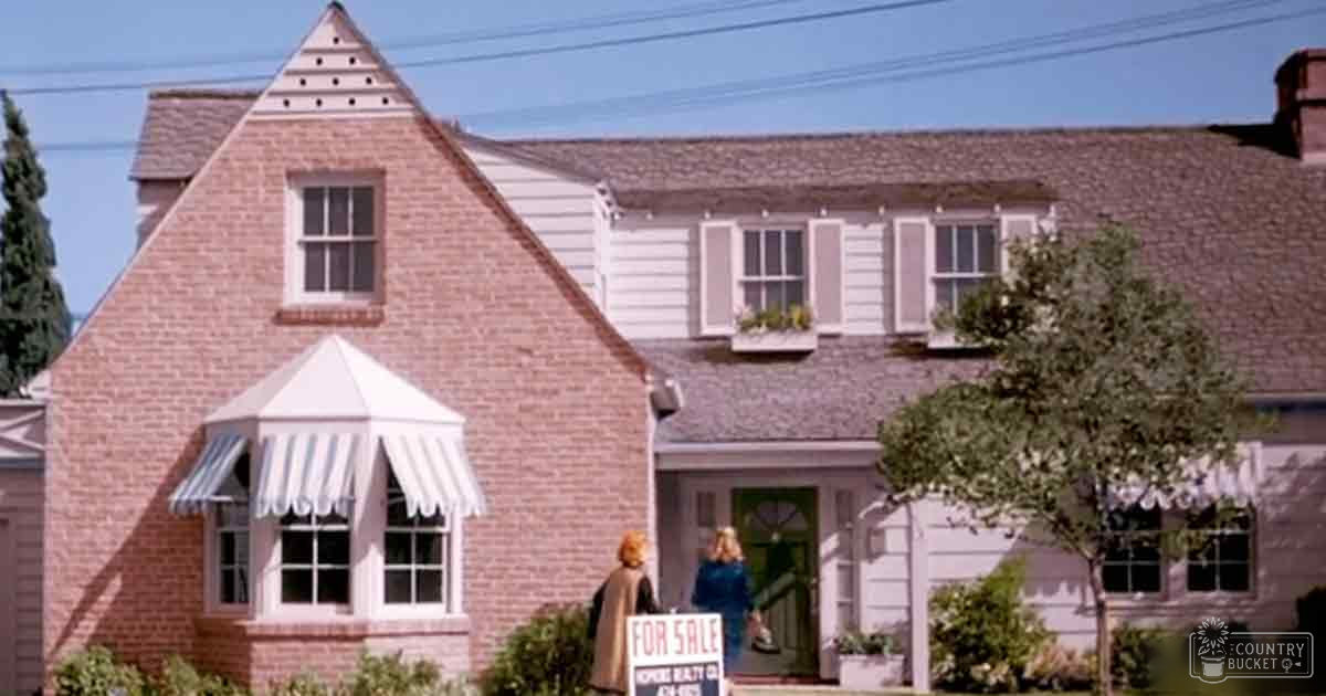 An adult female in a dark brown coat with long sleeves stands near a 'For Sale' sign, indicating a house for sale in an urban residential area. Another adult female in a blue coat is also present.