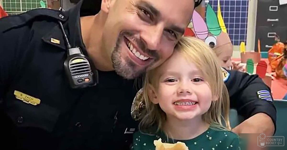 A young adult male police officer wearing a dark uniform shirt with patches, interacting with a toddler in a green dress with small white dots at what appears to be an indoor community event setting.