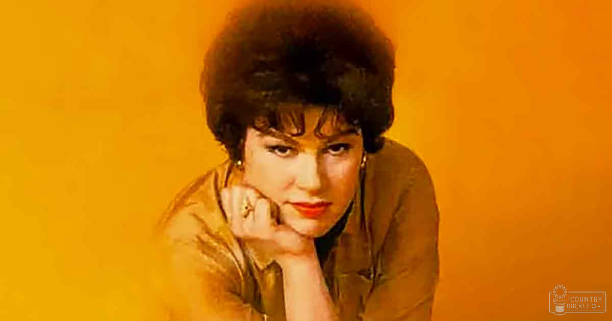 A young adult female wearing a short-sleeved, fitted blouse with a rounded neckline, likely from the mid-20th century, indoors against an orange background.