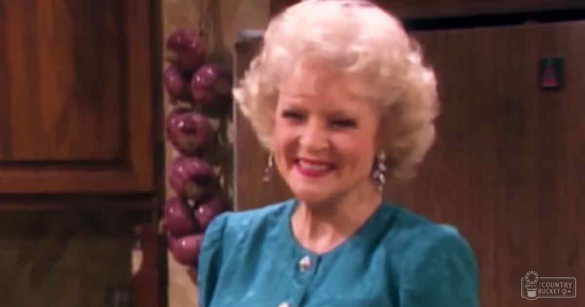 An older adult woman in a teal blouse stands in an indoor kitchen setting with natural lighting and visible wooden furniture.