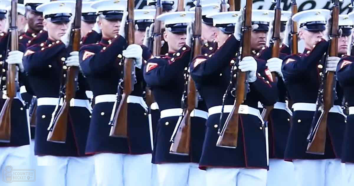 A group of young adult male military personnel in formal dark blue dress uniforms with white trim and buttons stand in formation outdoors, holding rifles vertically at the shoulder under a clear sky with sunlight illuminating the scene.