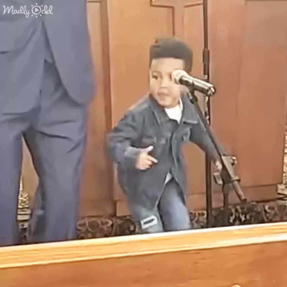 A young male child wearing a dark blue jacket and light-colored pants performs indoors at what appears to be a church setting with wooden pews visible.