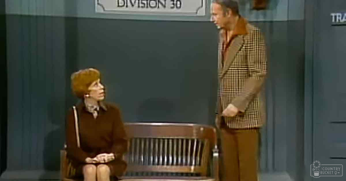 An adult female seated in Division 30, wearing a brown collared dress with buttons down the front. An adult male stands nearby in a checkered blazer and collared shirt.