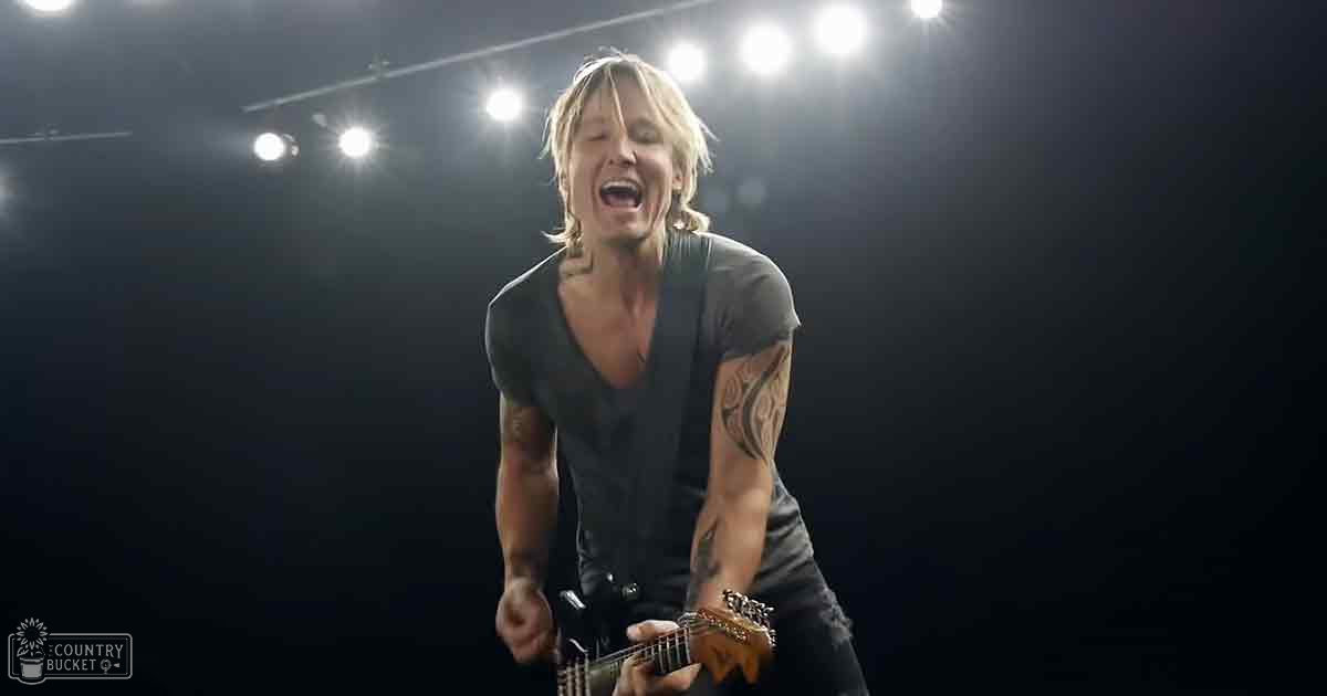 An adult male singer performing on stage, wearing a dark gray V-neck t-shirt with tattoos and earrings, holding a guitar under bright stage lights.