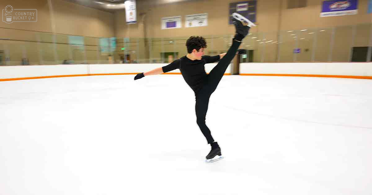 A young adult male figure skater in a black long-sleeve leotard and tights, skating indoors at an ice rink with glass walls and banners overhead.