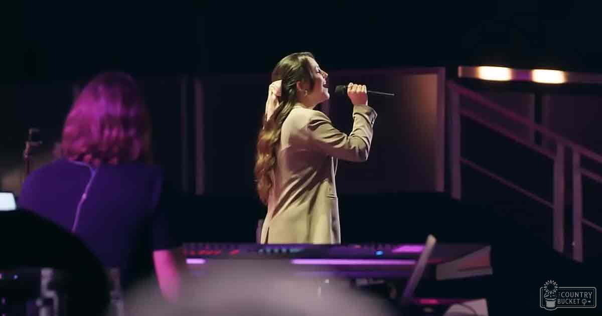 A young adult female singer performs on stage, wearing a light beige or tan blazer over a darker top at an indoor concert setting with purple and blue stage lighting.