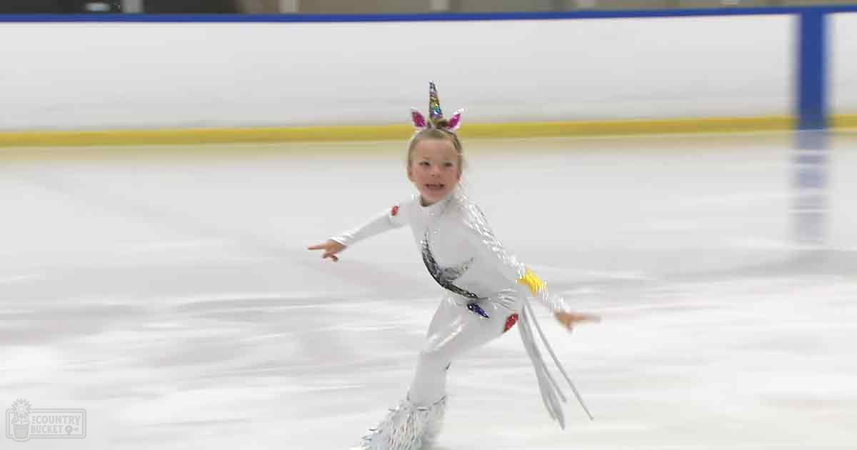 A toddler girl wearing a sparkly white leotard with black accents, skating on an indoor ice rink with blue barriers.