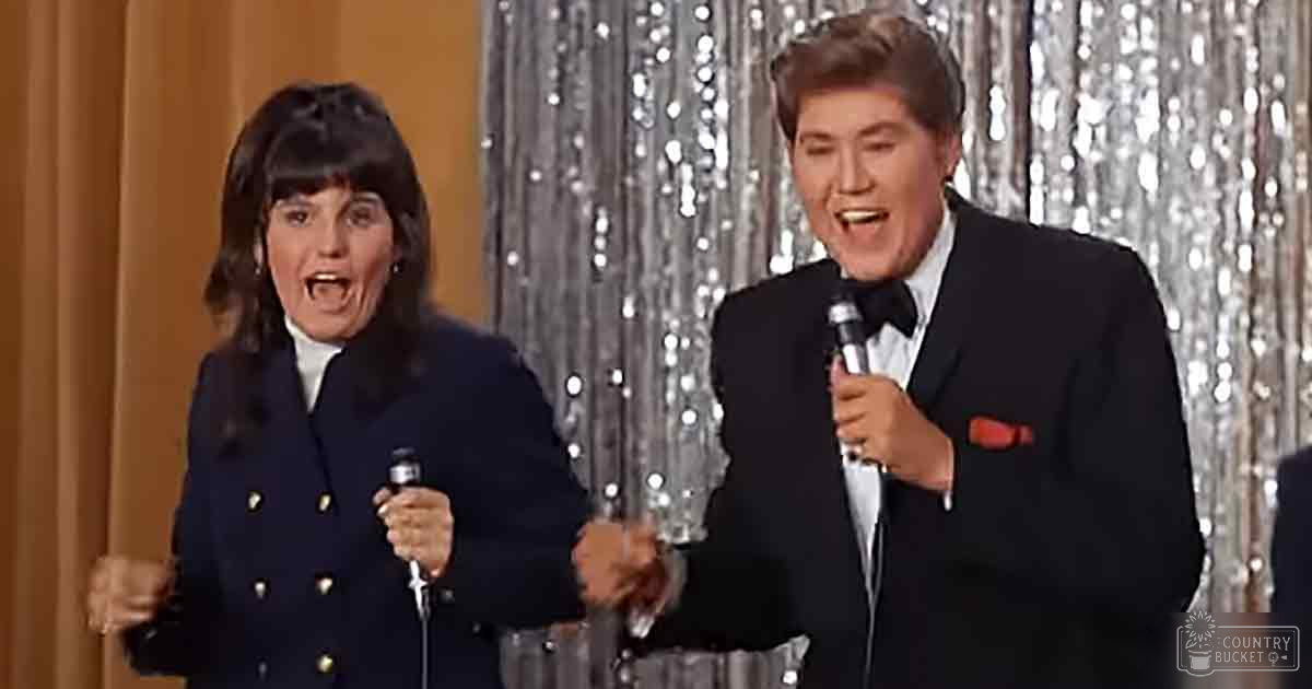 Adult female wearing a dark blue double-breasted jacket with gold buttons and white trim, alongside an adult male in a black tuxedo with a white shirt, black bow tie, and red pocket square. Microphones are held by each person on stage behind reflective silver sequin curtains.