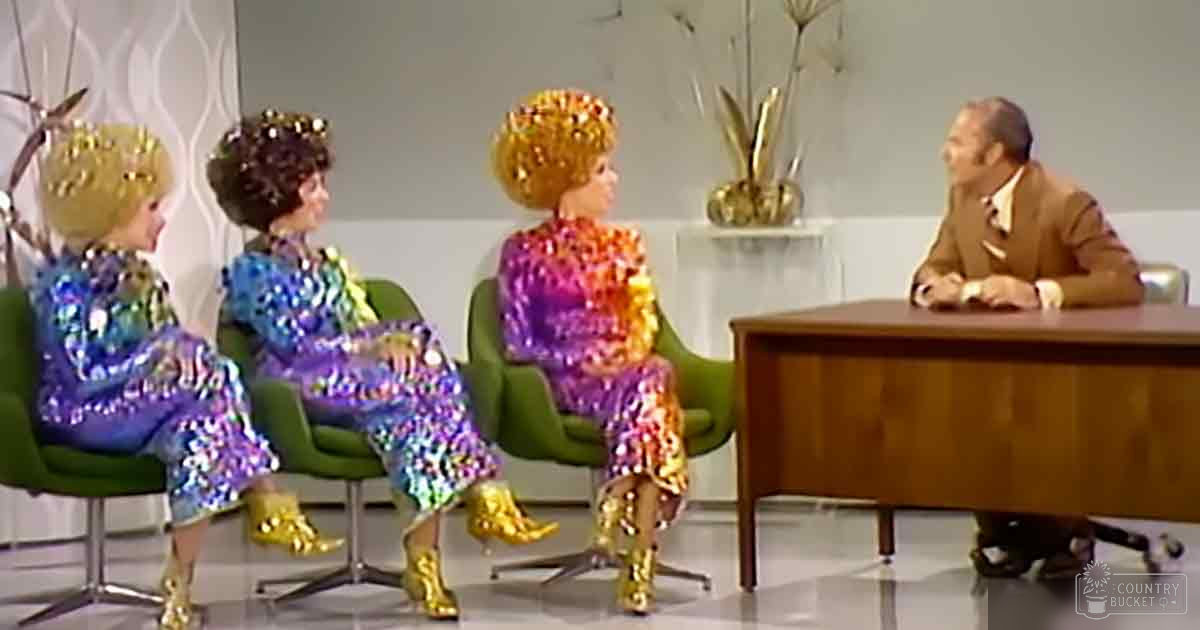 Talk show set with three adult females in shiny, sequined dresses and an adult male host in formal attire; vibrant, mid-20th century studio with green armchairs and a decorative vase.
