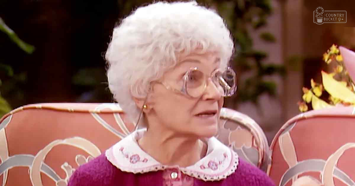 An older adult female in a cozy domestic setting, wearing a purple floral embroidered sweater with glasses and small earrings.