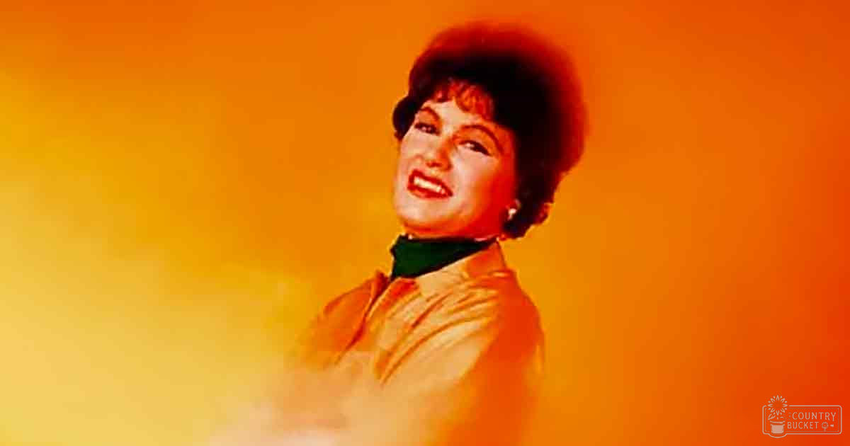 Middle-aged female in a light-colored blouse with a high neckline, set against an orange gradient background.