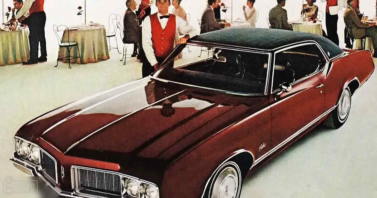 An Oldsmobile Cutlass displayed in a showroom with a waiter wearing formal attire and other adults dining in the background, suggesting an exhibition setting from the mid-20th century.