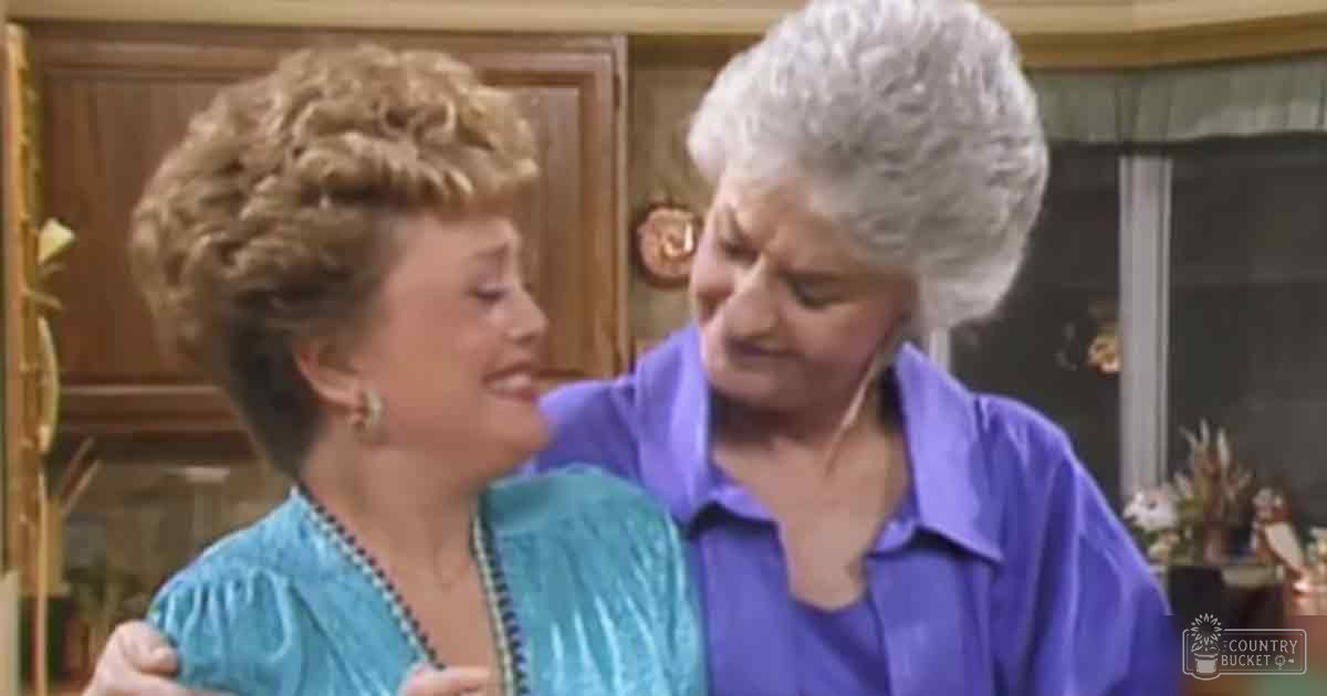 Two older adults, one in a teal blue textured dress and the other in a purple blouse, indoors with wooden furniture and decorative items.