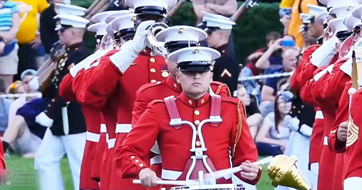 A marching band performance at an outdoor event with young adult males in red military-style uniforms, surrounded by trees and a grassy field during the afternoon.