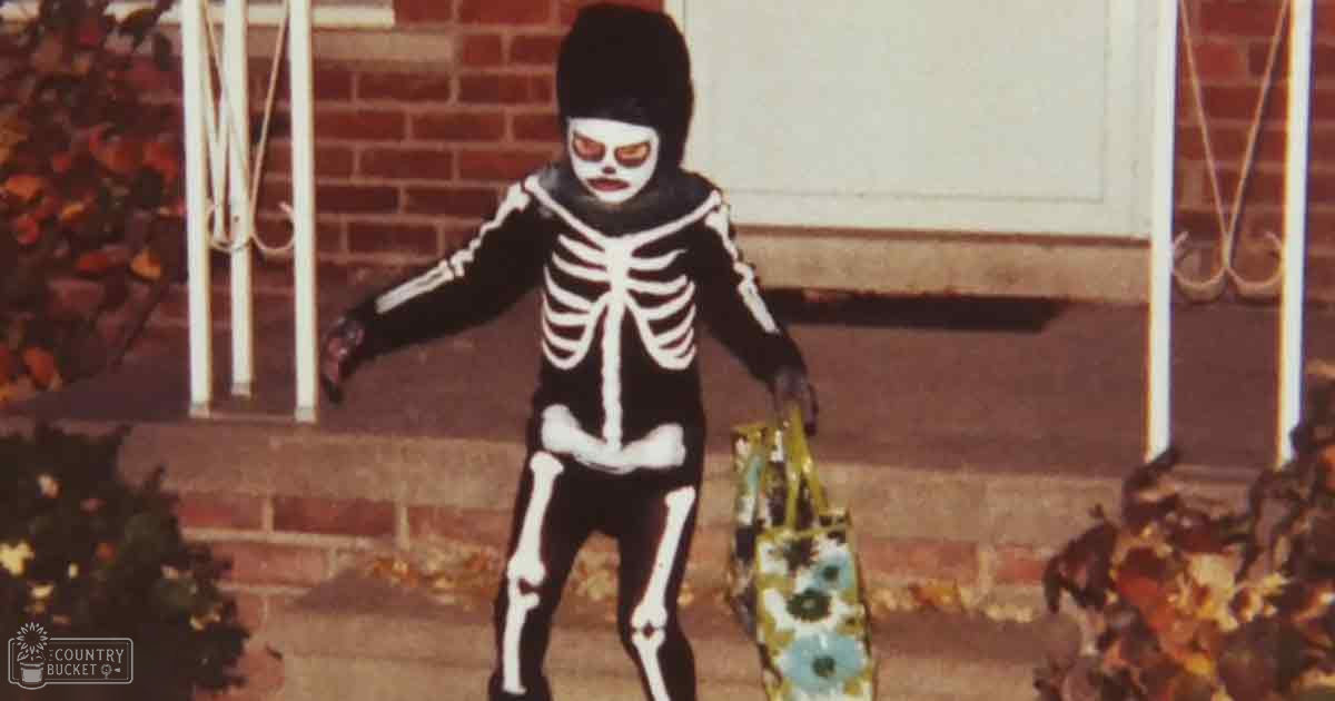 A toddler in a full-body skeleton costume near a house, holding a yellow bag with blue floral patterns; outdoor residential area during fall or early winter.