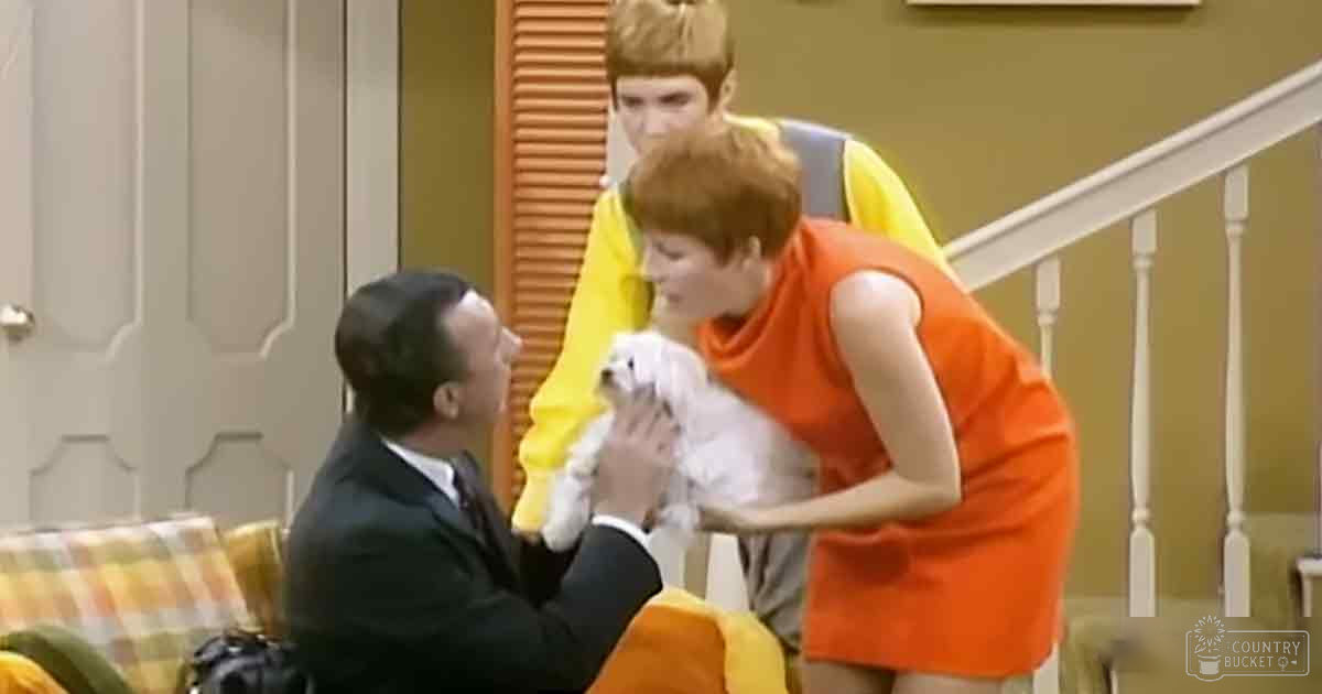 A man in a dark suit kneels in an indoor living room with a young adult male and a woman holding a small white dog. The setting includes a plaid-patterned couch, a staircase with white railings, yellow walls, and paneling on the door.