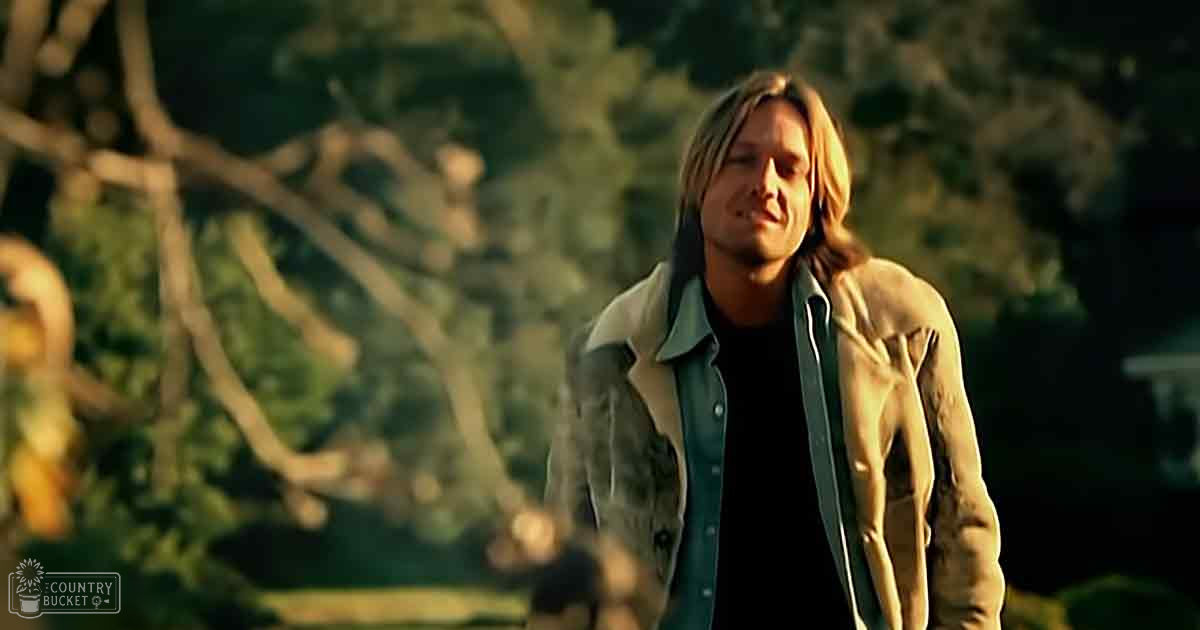A young adult male stands outdoors with trees and possibly a house visible in the background, wearing a light-colored jacket over a dark shirt. The scene is bathed in warm tones suggesting early morning or late afternoon.