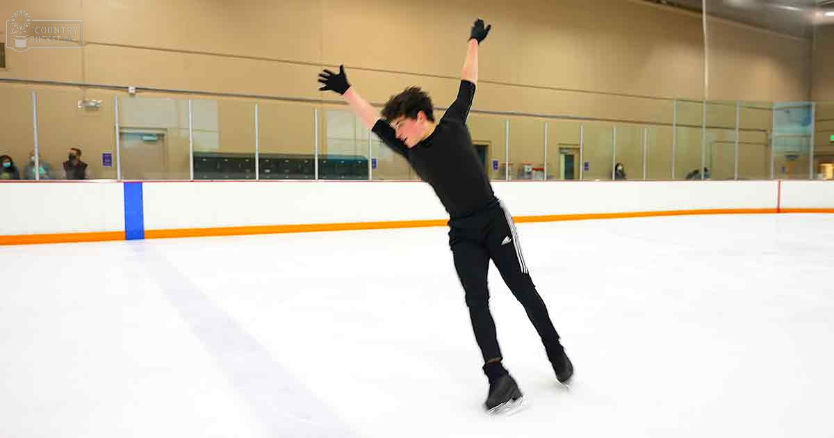 A young adult male figure skating in an indoor ice rink with a glass barrier, wearing black attire and gloves.