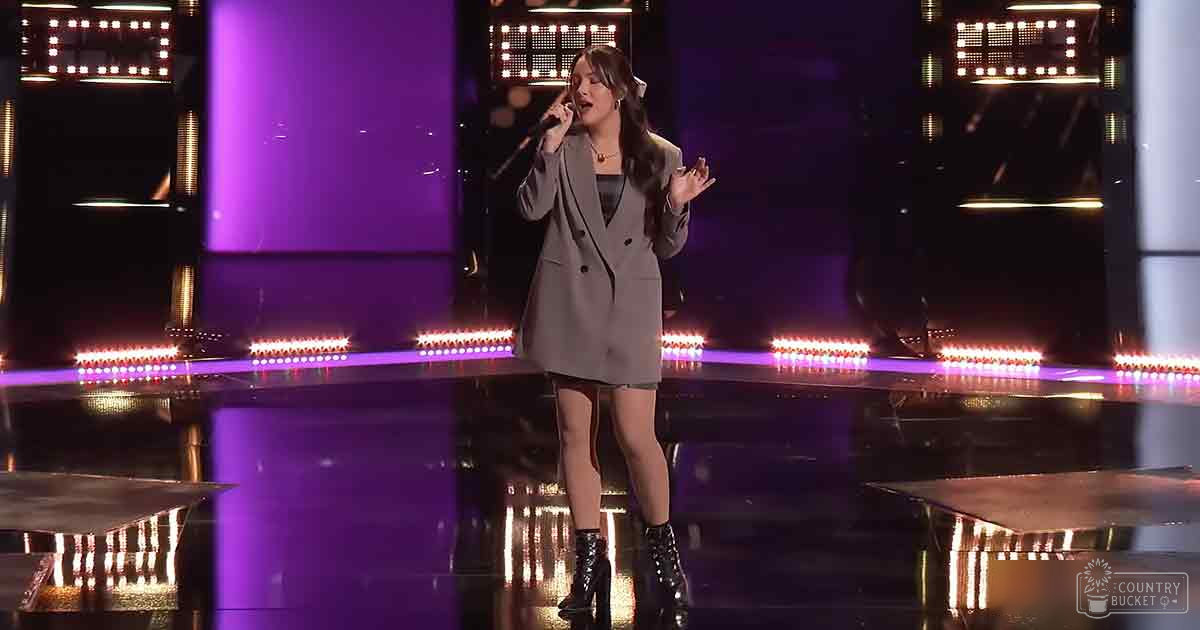 A young adult female in a double-breasted blazer dress with long sleeves and high-heeled boots performs on stage under purple and white lighting.