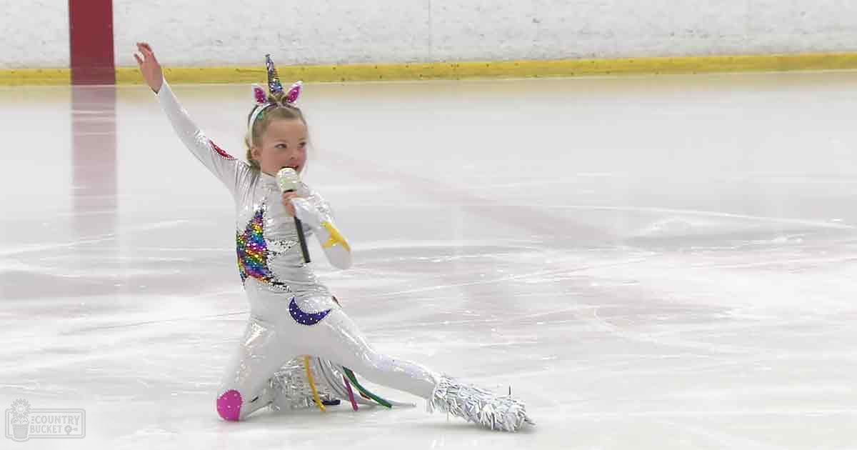 A toddler in a sparkly silver leotard with colorful unicorn designs, wearing pink ice skates and a unicorn headband, skating on an indoor ice rink.