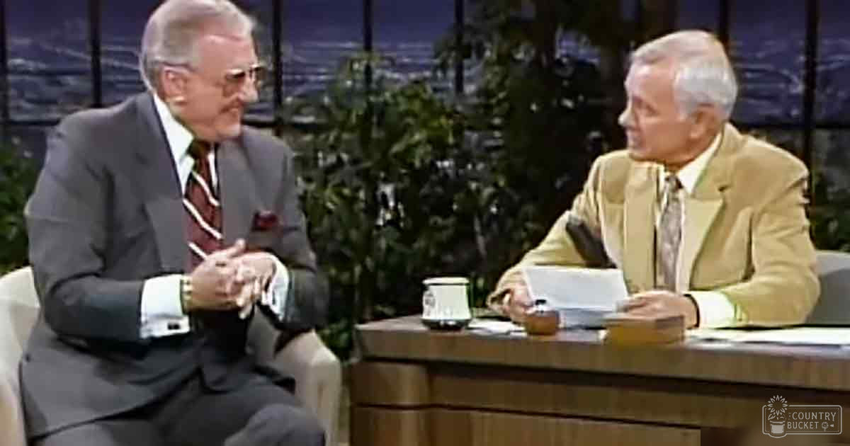 An older adult male talk show host and guest dressed formally, seated at a desk with cityscape in background.