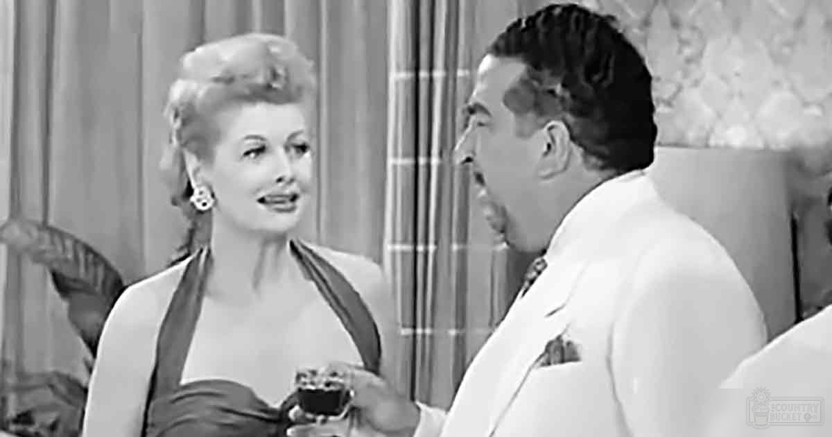 A woman wearing a strapless dress with wide straps stands indoors near an adult male in a white suit jacket. The setting suggests a bedroom, softly lit and likely from the mid-20th century.