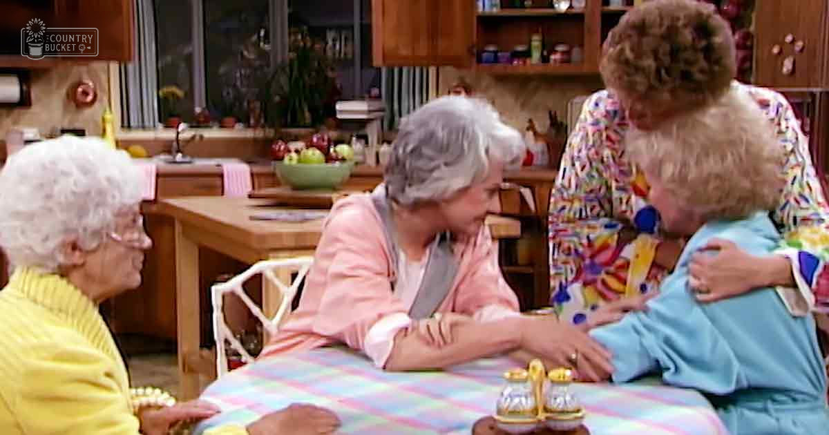A group of older adult females in a kitchen setting with wooden furniture and pastel-colored decor.